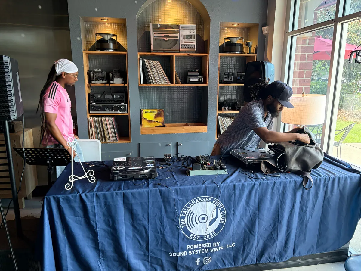 Two DJs at the club table, shelves of records, cassettes and vintage stereo gear behind them